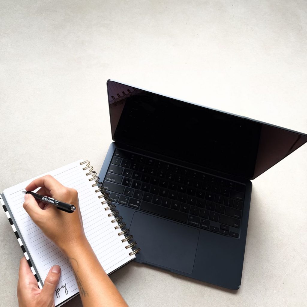 Manos escribiendo en una libreta junto a una laptop, categoría Cómo Vivo Mi Estilo